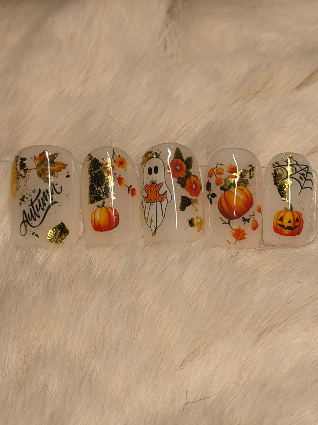 Five clear press-on nails with autumn and Halloween designs, including pumpkins, a ghost, flowers, leaves, spiderwebs, and gold foil accents, displayed on a soft beige faux fur background.