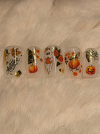 Five clear press-on nails with autumn and Halloween designs, including pumpkins, a ghost, flowers, leaves, spiderwebs, and gold foil accents, displayed on a soft beige faux fur background.