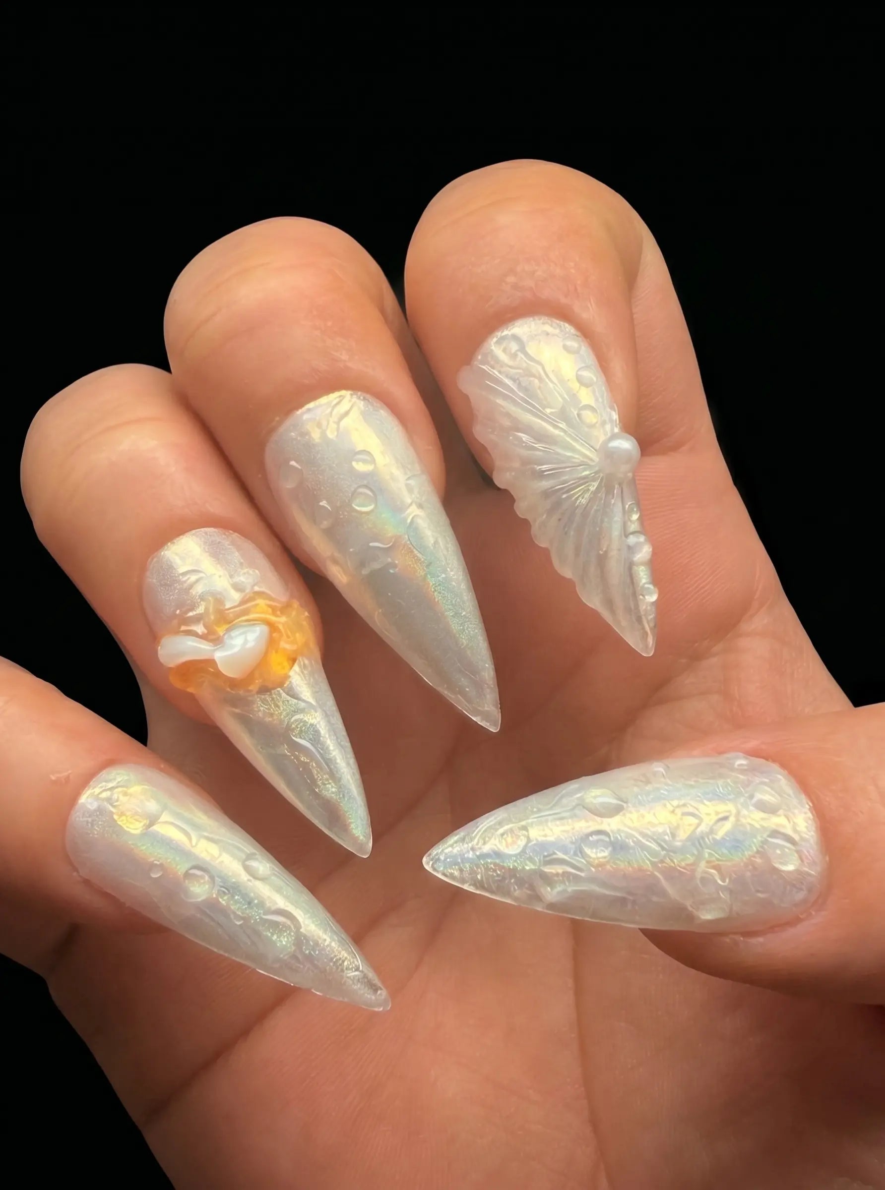 A close-up of a hand showcasing long, stiletto-shaped press-on nails with a glossy, iridescent white finish. One nail features a 3D white butterfly design, another has a white pearl accent, and one nail displays a delicate white floral pattern with a small orange accent.