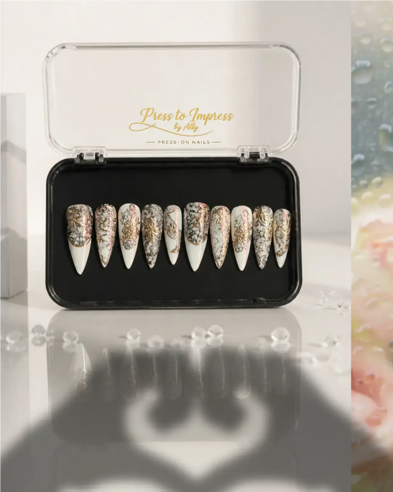 Handmade luxury press-on nails featuring pearl white chrome, black lace nail art, metallic gold floral details, glossy finish, and long stiletto shape.