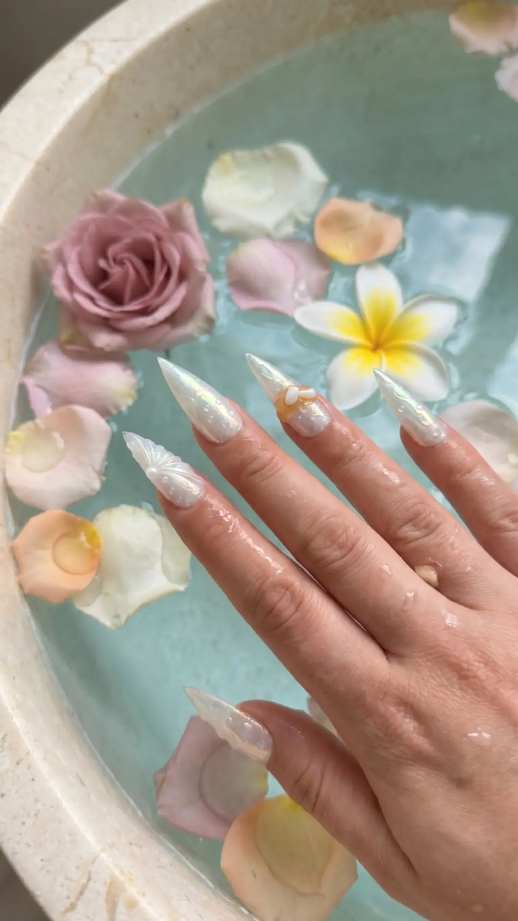 Luxury stiletto press-on nails featuring iridescent pearl chrome, realistic water droplet effects, translucent sculptural details, amber glass accents, and pearl embellishments.