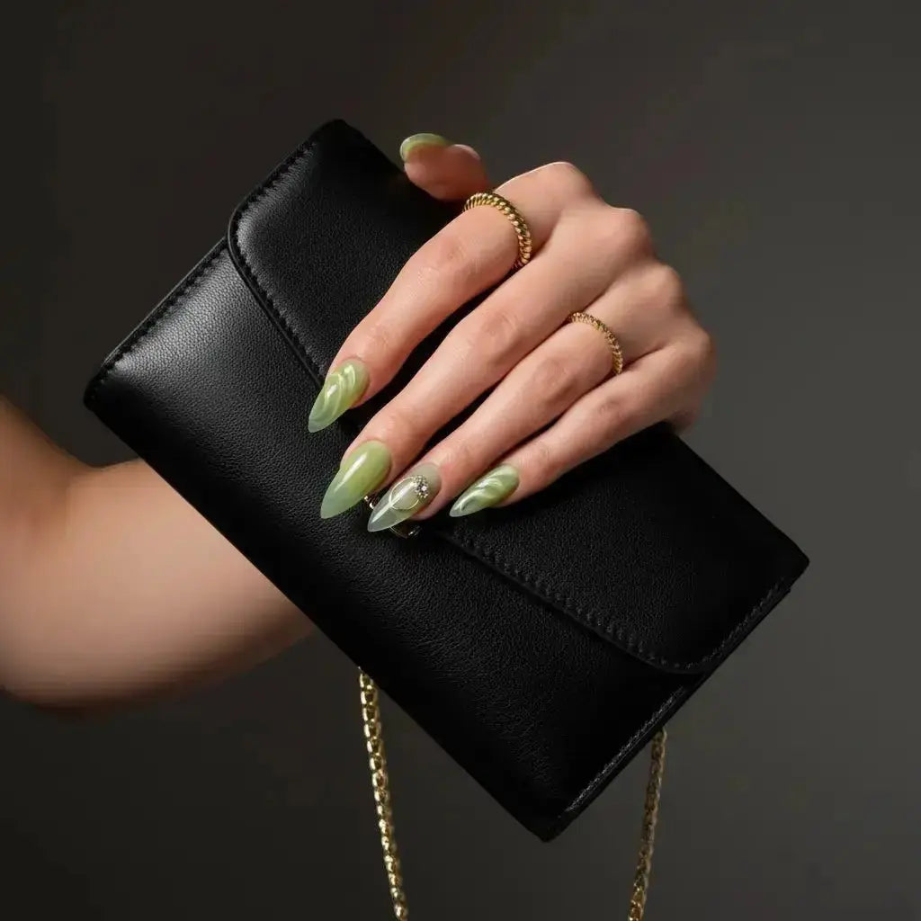 Press to Impress Pistachio Veil: A hand with long, stiletto nails in swirled pistachio and translucent green, adorned with rings, holds a black leather clutch with a gold chain.